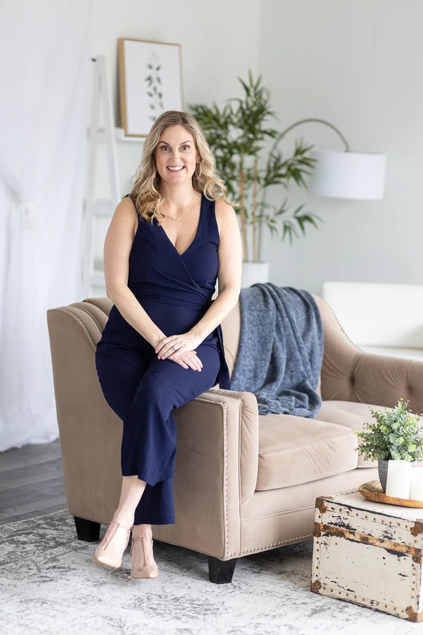 Smiling woman in navy jumpsuit sitting on beige chair in bright, modern skincare studio.